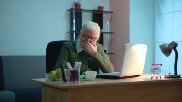 Пожилая женщина кашляет, может быть, она больна, сидит за столом, работает за компьютером — стоковое видео