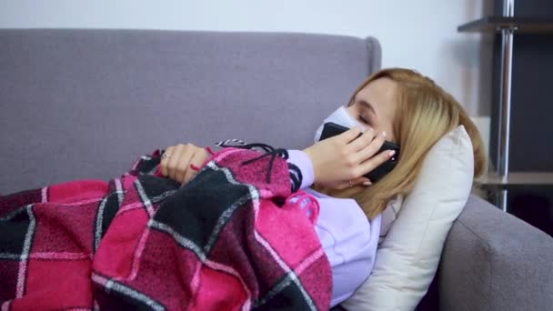 Chica acostada en un sofá con una máscara protectora cubierta con una manta hablando por teléfono, casa, enferma — Vídeos de Stock