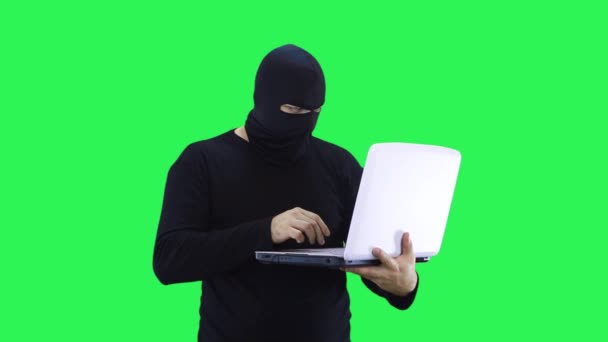 Een man met een zwart masker voert data in op een laptop, op een groene achtergrond — Stockvideo