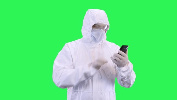 Un homme portant un masque et une combinaison de protection parle par vidéoconférence sur un smartphone tout en se tenant debout sur un fond vert . — Video