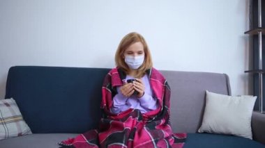 Kız hasta, elinde koruyucu bir maskeyle kanepede oturuyor, elinde bir fincan var, koronavirüs sıcaklığında.