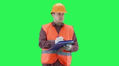 İnşaat kaskı ve turuncu yelek giyen bir adam verileri kağıda yazıyor, yeşil ekran arka planı.