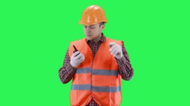 İnşaat kasklı ve turuncu yelekli bir adam yeşil ekran arka planında telsizle konuşuyor.