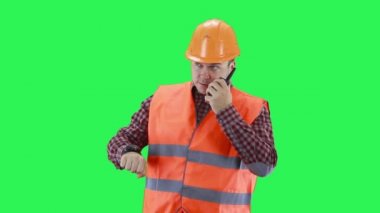 İnşaat kasklı ve turuncu yelekli bir adam saatine bakıyor, telsizle konuşuyor, yeşil ekran arka planında.
