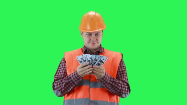Человек в строительном шлеме и оранжевом жилете пересчитывает деньги, зеленый фон — стоковое видео
