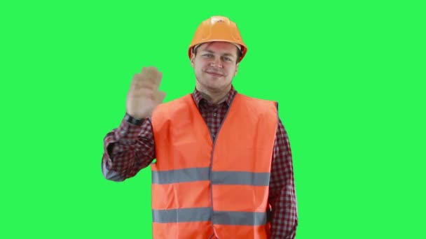 Un hombre en un casco de construcción y un chaleco naranja sonrisas y olas, fondo de pantalla verde — Vídeos de Stock