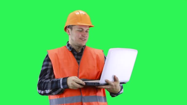 Un hombre en un casco de construcción y un chaleco naranja está hablando en un ordenador portátil en un buen estado de ánimo, fondo de pantalla verde — Vídeo de stock