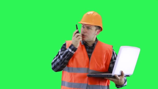 En man i bygghjälm och orange väst pratar på en walkie-talkie och kollar data på en bärbar dator. — Stockvideo