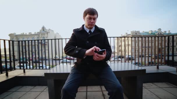 검은 코트를 입고 벤치에 앉아 있는 사업가가 전화로 스마트폰을 하고 있습니다. — 비디오