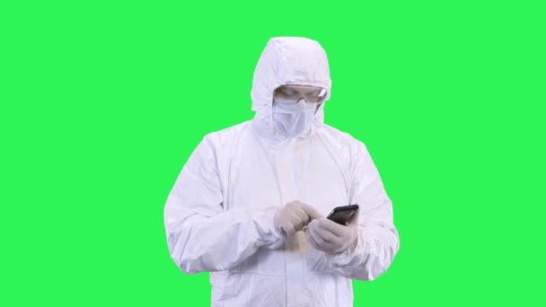 Человек в маске в защитном костюме сёрфинг телефон, стоя на зеленом фоне — стоковое видео