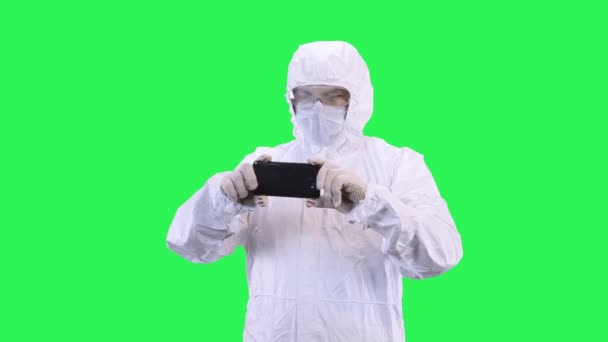 Een man met een masker en een beschermend pak schiet op een smartphone terwijl hij tegen een groene achtergrond staat. — Stockvideo