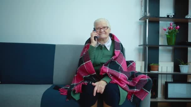 Бабушка разговаривает по телефону, завернутая в клетчатую клетку, сидя на диване — стоковое видео