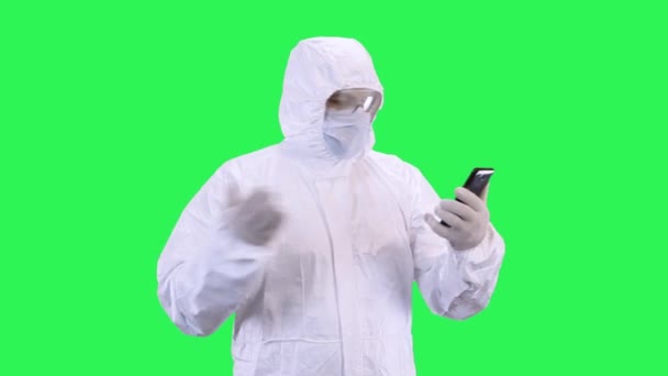 Un hombre con una máscara y un traje protector está hablando a través de videollamadas en un teléfono inteligente mientras está de pie sobre un fondo verde, peleando y maldiciendo con un interlocutor — Vídeos de Stock