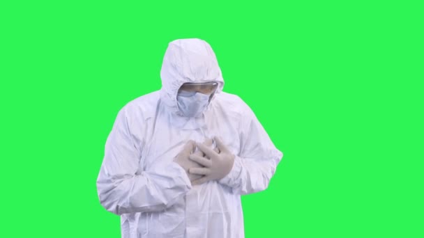 Ein Mann im Schutzanzug mit Maske auf dem Kopf und Brille hält sich an einem Herzen auf grünem Hintergrund fest — Stockvideo