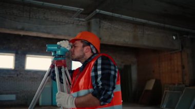 Turuncu yelek ve turuncu kasklı bir adam, bir inşaat alanının arka planındaki ölçüm cihazına bakıyor..