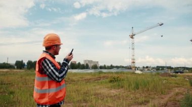 Turuncu yelekli ve miğferli bir adam, bir telsize doğru konuşur, kuleye bakar ve elleriyle ne yapacağını gösterir..