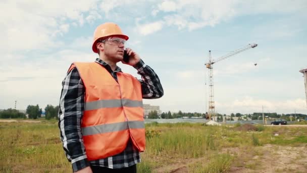En man i orange gult och en konstruktion hjälm står vid en byggarbetsplats på bakgrunden av en kran, ser, samtal på telefonen. — Stockvideo