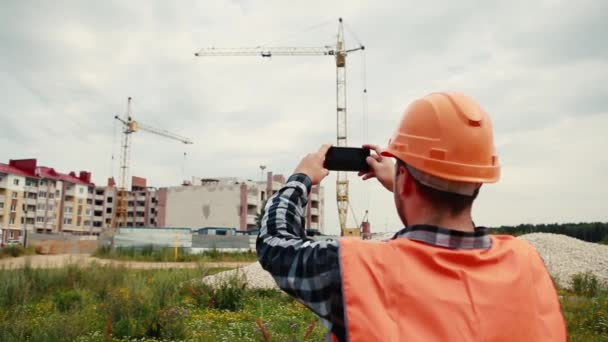 Un constructeur en gilet orange et un casque orange filment une vidéo de construction sur un téléphone portable . — Video