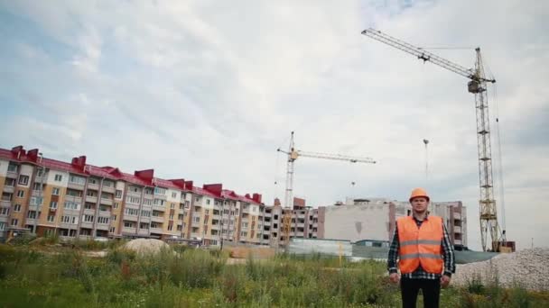 En byggare i orange väst och en orange hjälm sprider sina armar i olika riktningar mot bakgrund av en byggarbetsplats. — Stockvideo