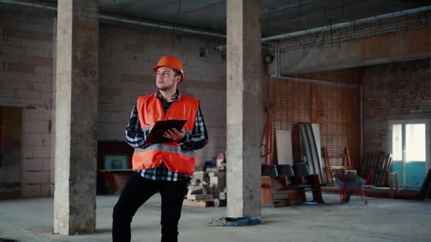 Een man in een oranje vest en een oranje helm staat tegen de achtergrond van een bouwplaats. — Stockvideo