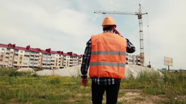 Le constructeur dans un casque et dans un gilet va à la grue à tour parler au téléphone . — Video