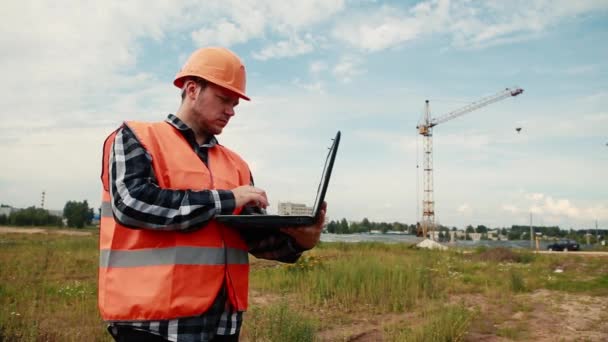 O construtor em um canteiro de obras no fundo de um guindaste entra em dados em um laptop . — Vídeo de Stock