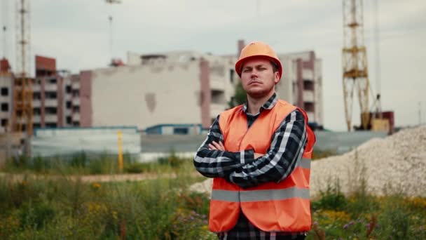 Een videoportret van een man in een oranje vest en een oranje helm die tegen de achtergrond van een bouwplaats staat. — Stockvideo