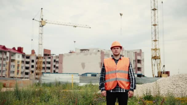 Un hombre con chaleco naranja y casco naranja extiende sus brazos en diferentes direcciones contra el telón de fondo de una obra de construcción . — Vídeos de Stock