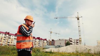 İnşaatçı inşaat alanının arka planında telsizle konuşuyor ve inşaat vincine bakıyor..