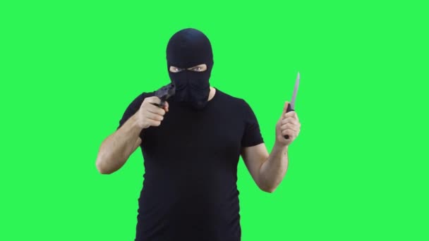 Un hombre con una máscara negra sostiene un cuchillo y un arma en su mano.Balaclava.Fondo de pantalla verde . — Vídeo de stock