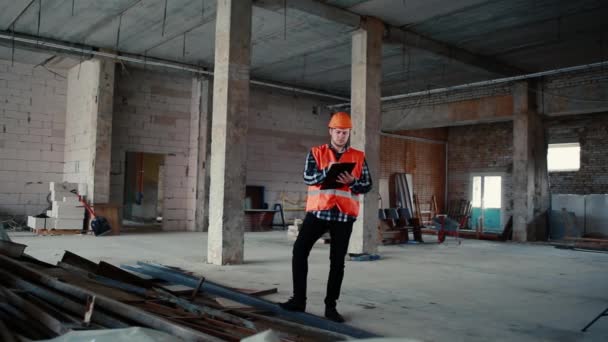 Un constructor con un chaleco naranja y un casco naranja se levanta contra el telón de fondo de una obra de construcción . — Vídeos de Stock