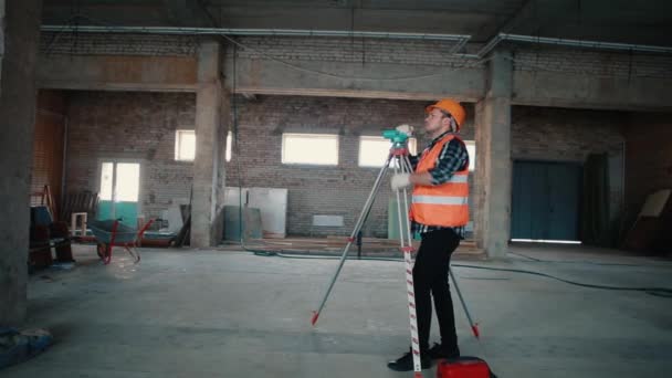 Un constructor con chaleco naranja y casco naranja está mirando un dispositivo de medición contra el telón de fondo de una obra de construcción . — Vídeo de stock