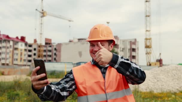 Un constructor con un chaleco naranja y casco naranja toma fotos de sí mismo en el fondo de una obra de construcción en un teléfono móvil . — Vídeo de stock