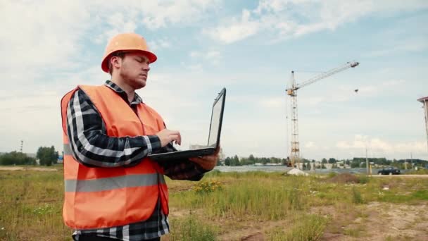 Un constructor en un sitio de construcción en el fondo de una grúa mira un objeto en construcción e ingresa datos en una computadora portátil . — Vídeo de stock