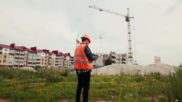 En byggare i en hjälm står med sin rygg skriva data mot bakgrund av ett hus under uppbyggnad. — Stockvideo