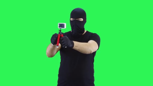 Человек в балаклаве фотографирует себя на экшн-камеру, зеленый фон — стоковое видео