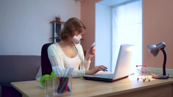 La chica trabaja en casa durante la cuarentena. pandemia de Coronavirus. Una chica con una máscara quirúrgica trabaja en una computadora portátil, ingresa datos de una tarjeta. Trabajo remoto . — Vídeos de Stock