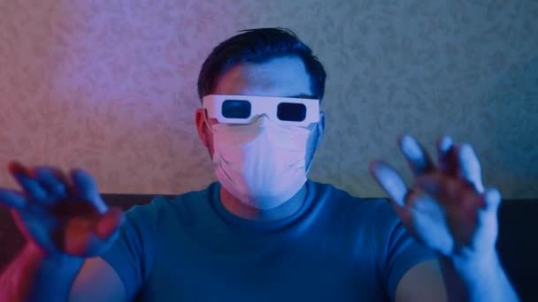 Um homem com uma máscara protetora e óculos estéreo assiste TV acenando com as mãos . — Vídeo de Stock