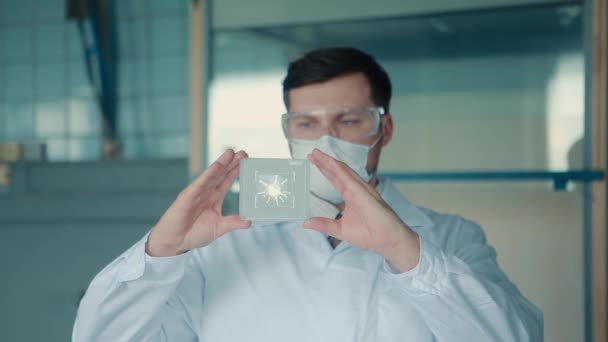 Um médico em uma máscara protetora segura em suas mãos um vidro médico virtual para análise em que o vírus é mostrado . — Vídeo de Stock