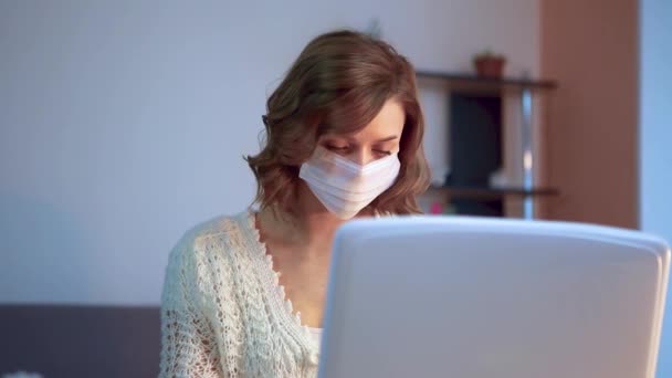 Flickan jobbar hemma under karantänen. En tjej i kirurgmask jobbar på en laptop. Fjärrarbete. — Stockvideo