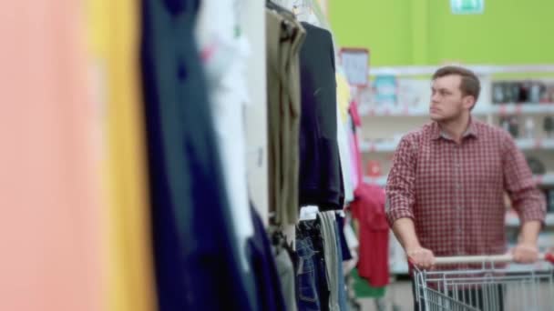 Un hombre con una camisa a cuadros selecciona las mercancías, mirando el estante selecciona una camiseta blanca y la pone en un carro . — Vídeo de stock