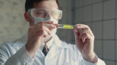 Koruyucu maskeli bir doktor elinde COVID-19 virüsü olan bir test tüpü tutuyor ve onu inceliyor. Virüslü bir test tüpünün yakın plan çekimi..