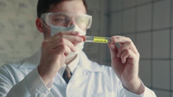Un médecin portant un masque de protection tient une éprouvette avec le virus COVID-19 dans les mains et l'examine. Gros plan d'une éprouvette contenant le virus . — Video