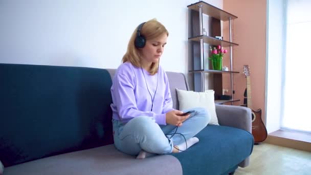 The girl on the couch and in the headphones sits on the couch listening to music and shakes her head. — Stock Video