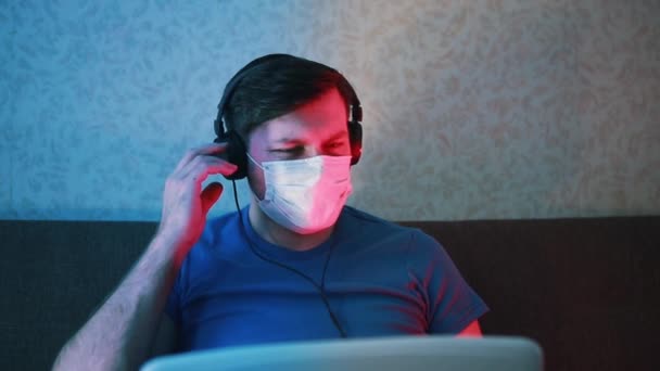 Человек в маске и наушниках работает на удаленном компьютере на ноутбуке . — стоковое видео