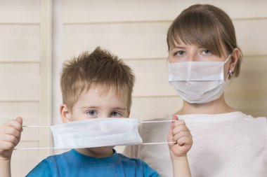 Çocuğun önünde tıbbi bir maske var. Maskeli annesi arkada.