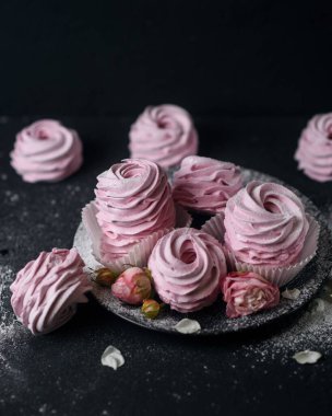 Ev yapımı tatlı, pembe marşmelov, kıvrımlı kağıtta zephyr, frenk üzümü aromalı, siyah bir tabakta, krema şekeri serpiştirilmiş, siyah arka planda güllerle süslenmiş, dikey fotoğraf, yakın çekim, yumuşak odak, ağız sulandırıcı fotoğraf