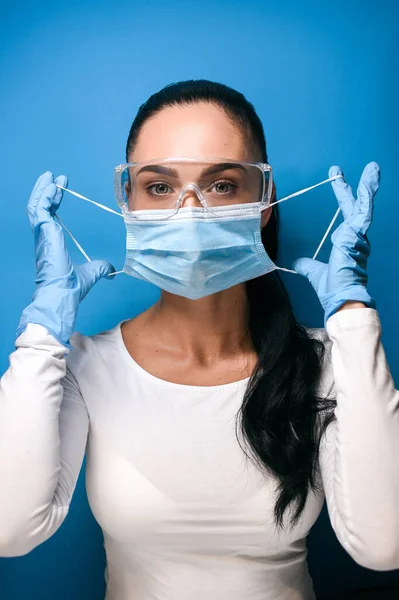 Young brunette girl with collected hair in green-eyed glasses, a protective medical mask, puts on a protective mask, in blue medical gloves, dressed in a white longsleeve, on a blue background, soft focus, vertical photo