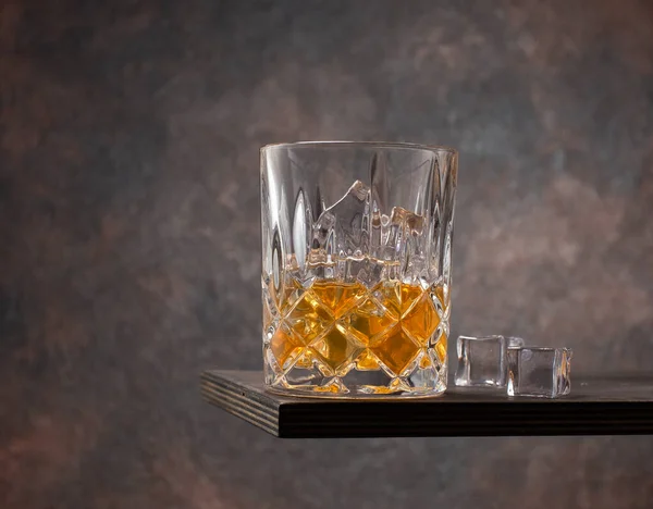 Scotch or whiskey in crystal glass with ice cubes on wooden table.
