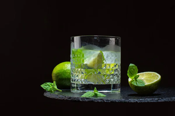 Cocktail freddo di mojito in cristallo o limonata di lime con lime, foglie di menta e cubetti di ghiaccio su lastra di pietra.Close up su sfondo scuro. — Foto Stock